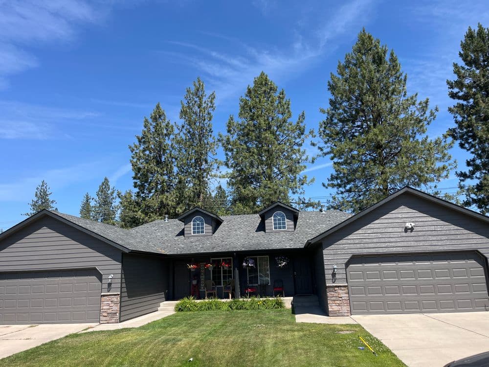Gray single-story house with two garages, surrounded by tall pine trees and a clear blue sky.