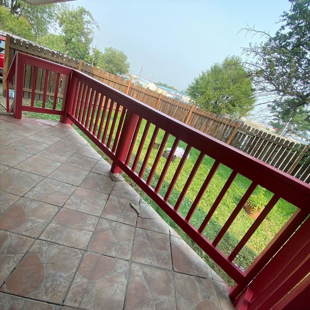 Red wooden railing on a patio overlooking a grassy yard and wooden fence.
