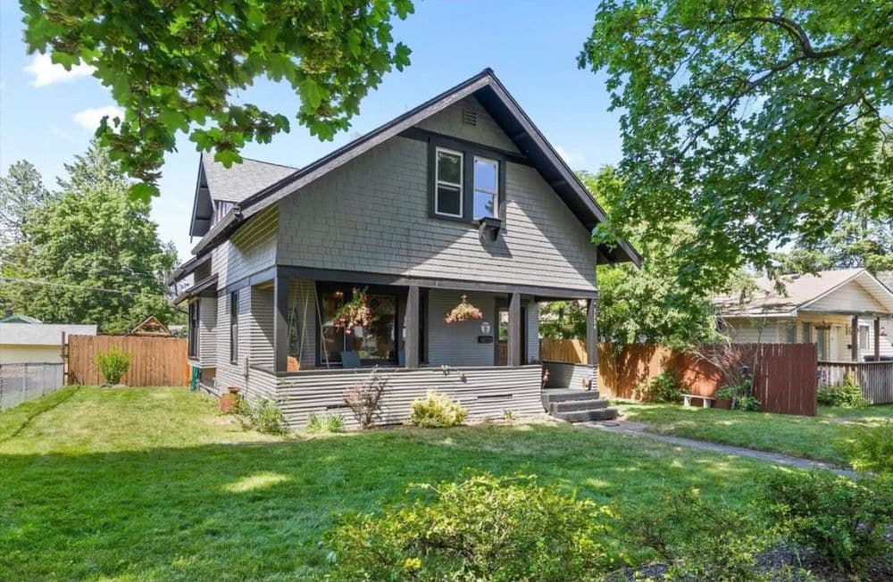 Two-story gray house with porch surrounded by green lawn and trees on a sunny day.