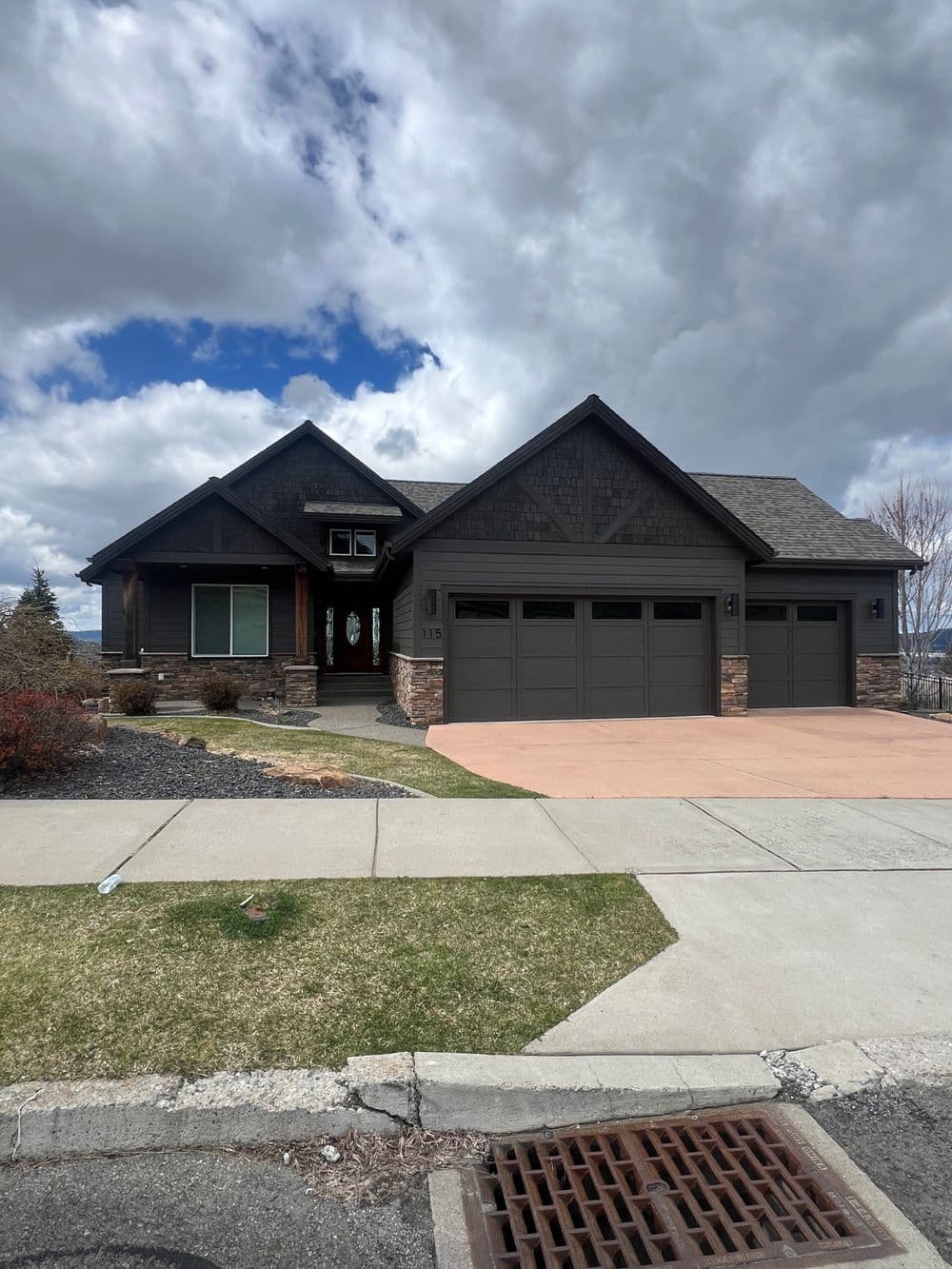 Modern dark house with stone accents, three-car garage, and landscaped front yard. Cloudy sky.