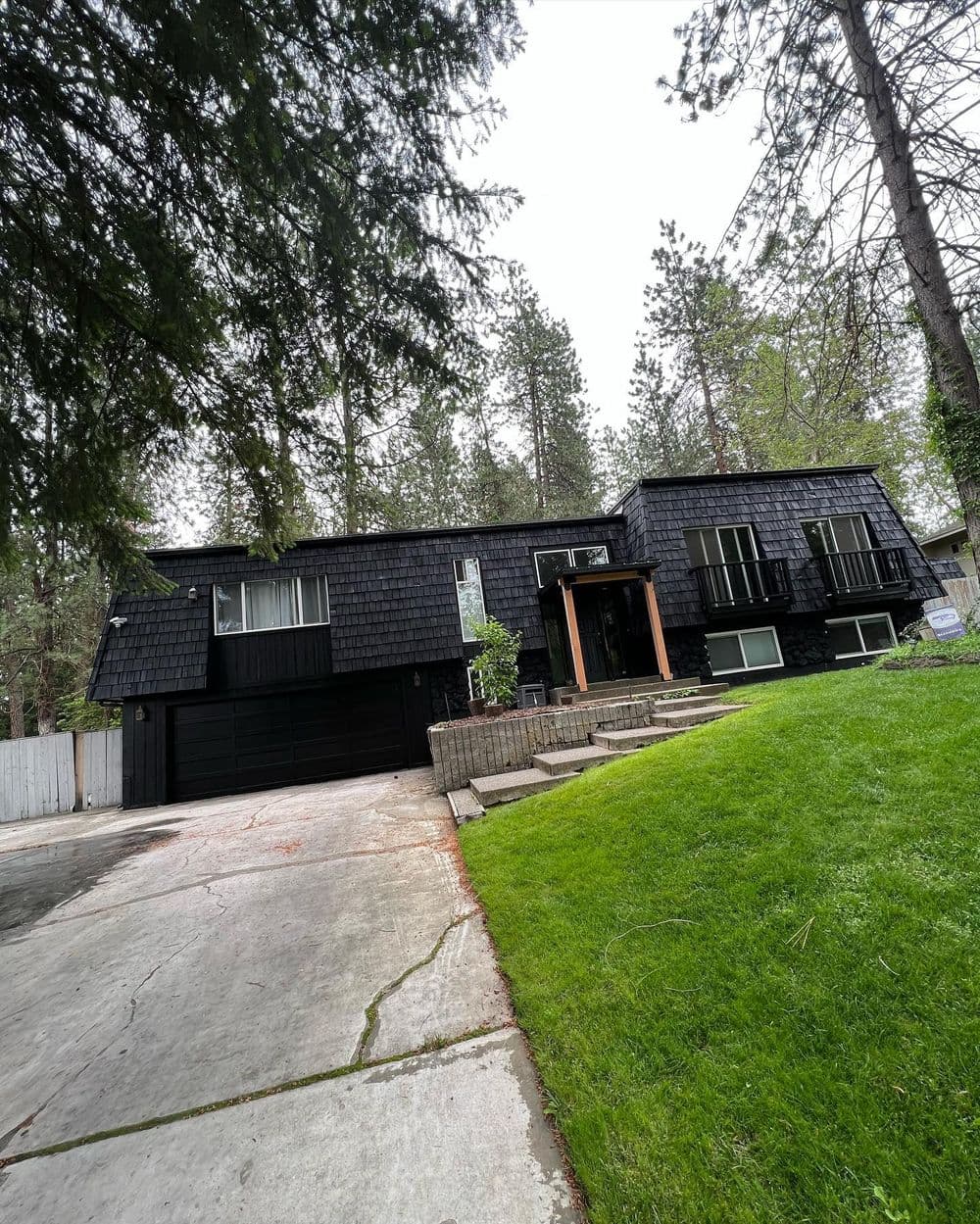 Modern black house with shingle siding, surrounded by greenery and a concrete driveway.
