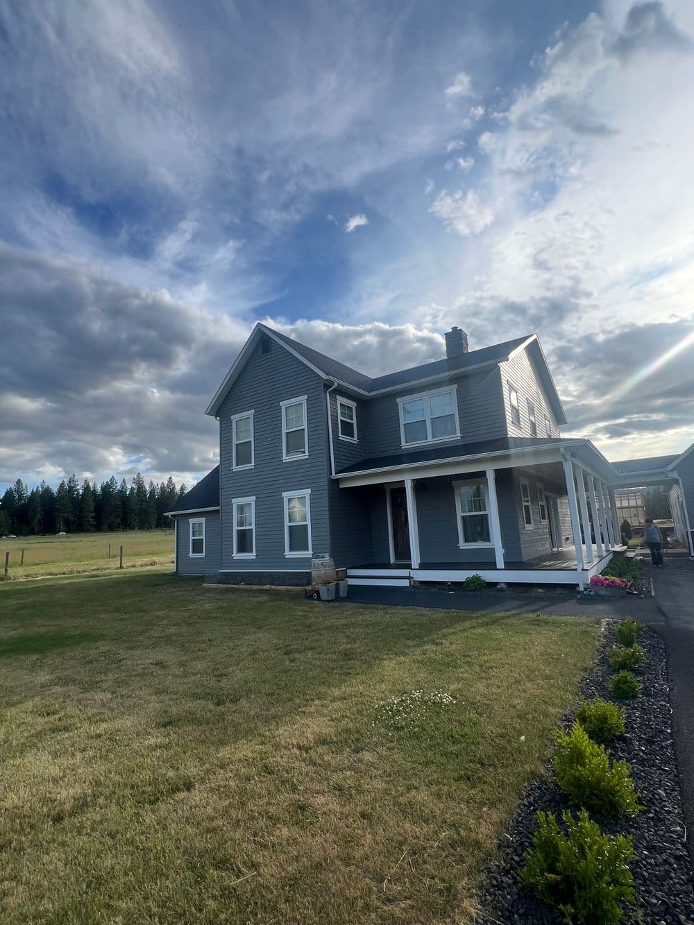 Gray two-story house with a porch, surrounded by a green lawn under a cloudy sky.