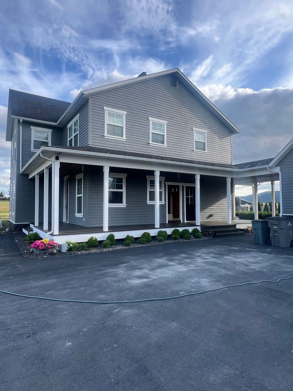 Gray two-story house with a porch, flower beds, and a driveway on a sunny day.
