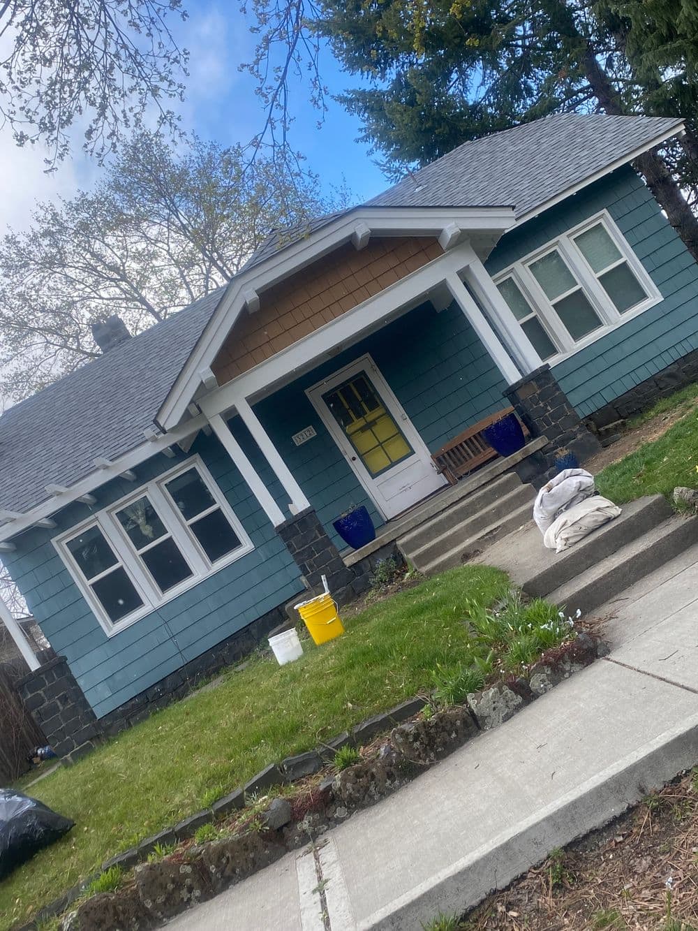 Charming blue bungalow with a porch, landscaped yard, and cloudy sky background.