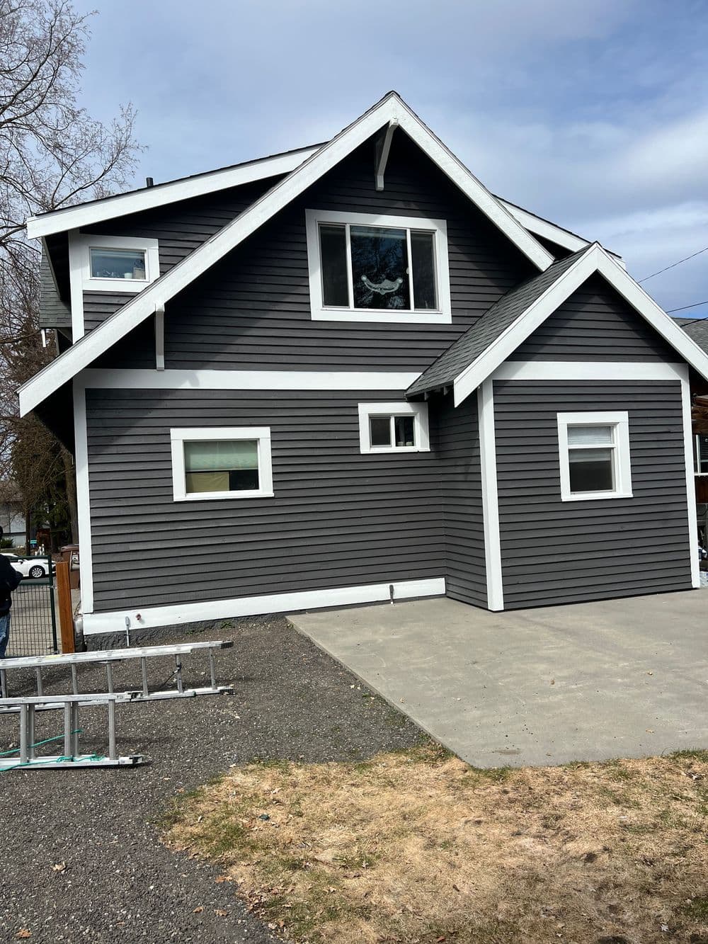 Modern gray house with white trim, driveway, and ladder in front yard under a cloudy sky.