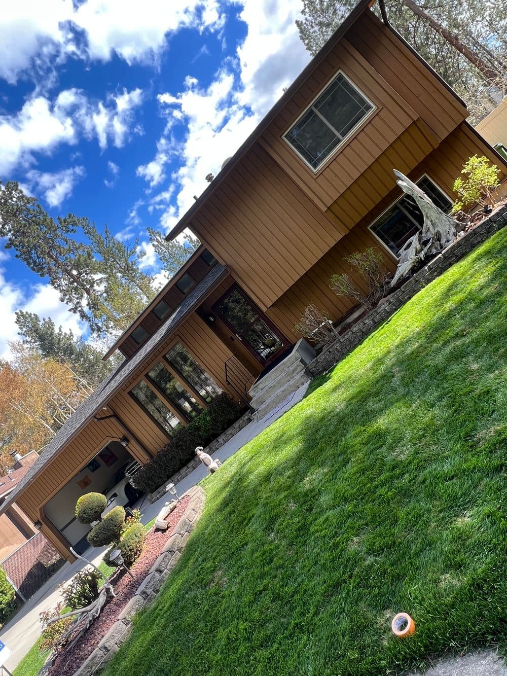Modern brown wooden house with green lawn and blue sky, surrounded by trees.