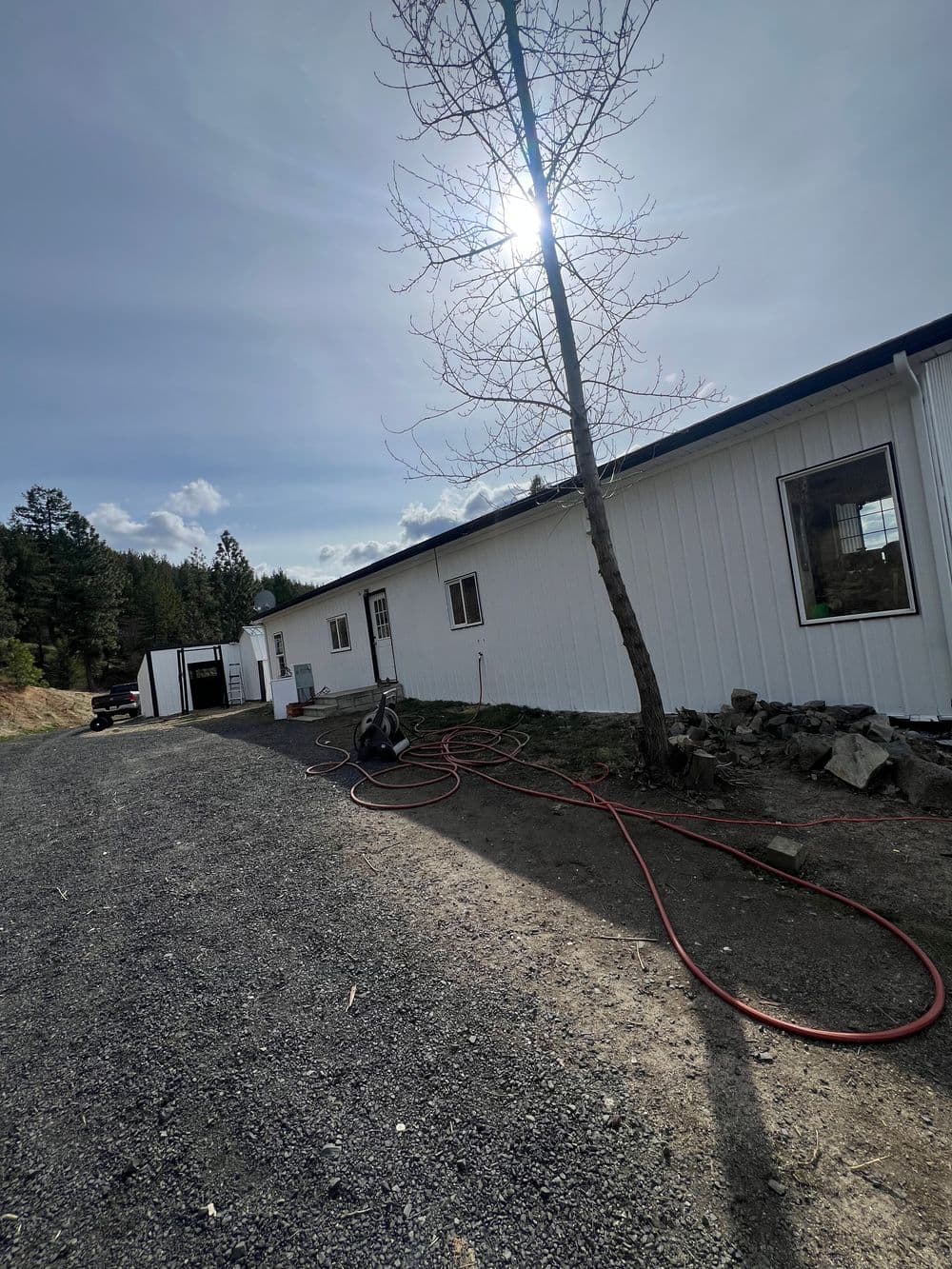 White building beside gravel path with tree and hose, surrounded by forested area.